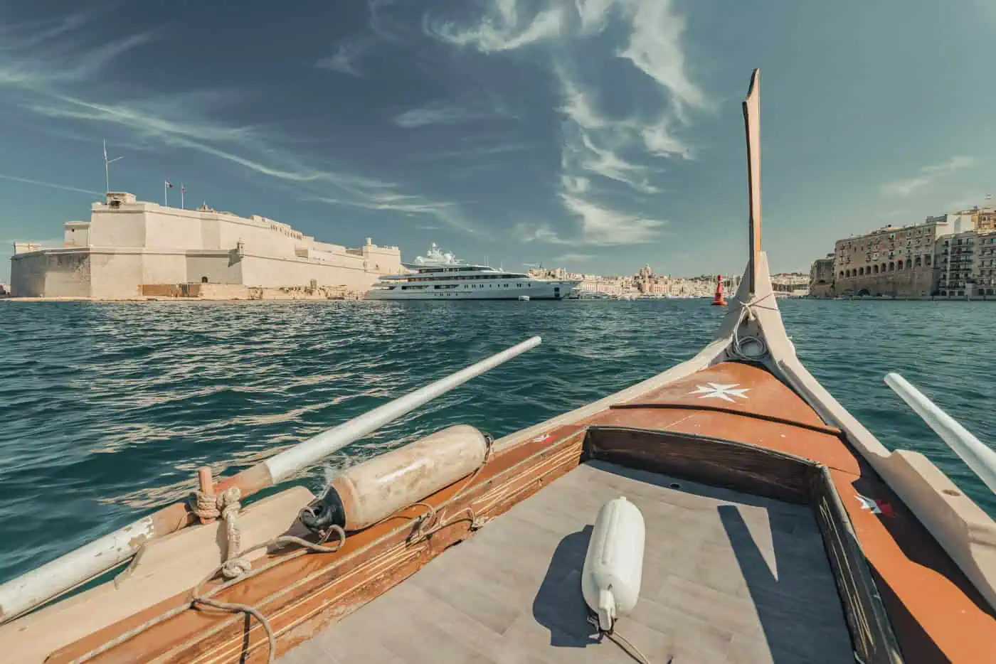 View from a wooden boat on turquoise water, with a large yacht, stone buildings, and a coastal cityscape in the background under a clear sky.