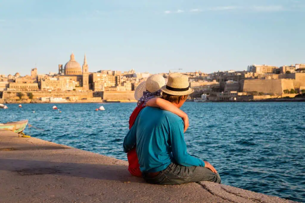 A couple sits on a waterfront promenade, facing a cityscape with historic buildings and an atmospheric church with a domed roof across the water in the afternoon light. They both wear hats, and one has an arm around the other.