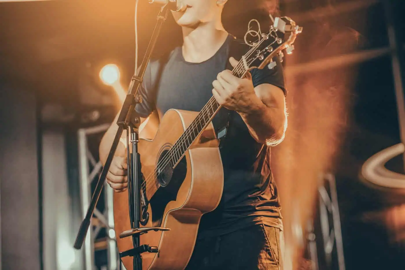 A person in a dark shirt plays an acoustic guitar, singing into a microphone on stage with warm lighting, capturing the soul of live music venues in Malta.