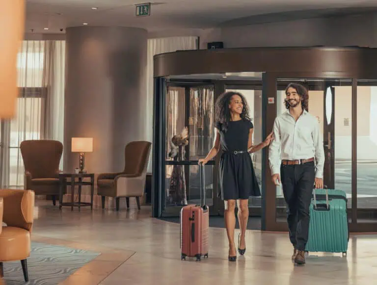 A man and woman, both casually dressed, walk arm in arm through a hotel lobby, each carrying a rolling suitcase.