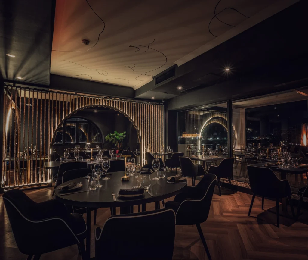 A dimly lit, modern restaurant interior with elegant round tables, neatly arranged place settings, and ambient lighting creating a cozy atmosphere. Large windows reveal a nighttime cityscape outside.