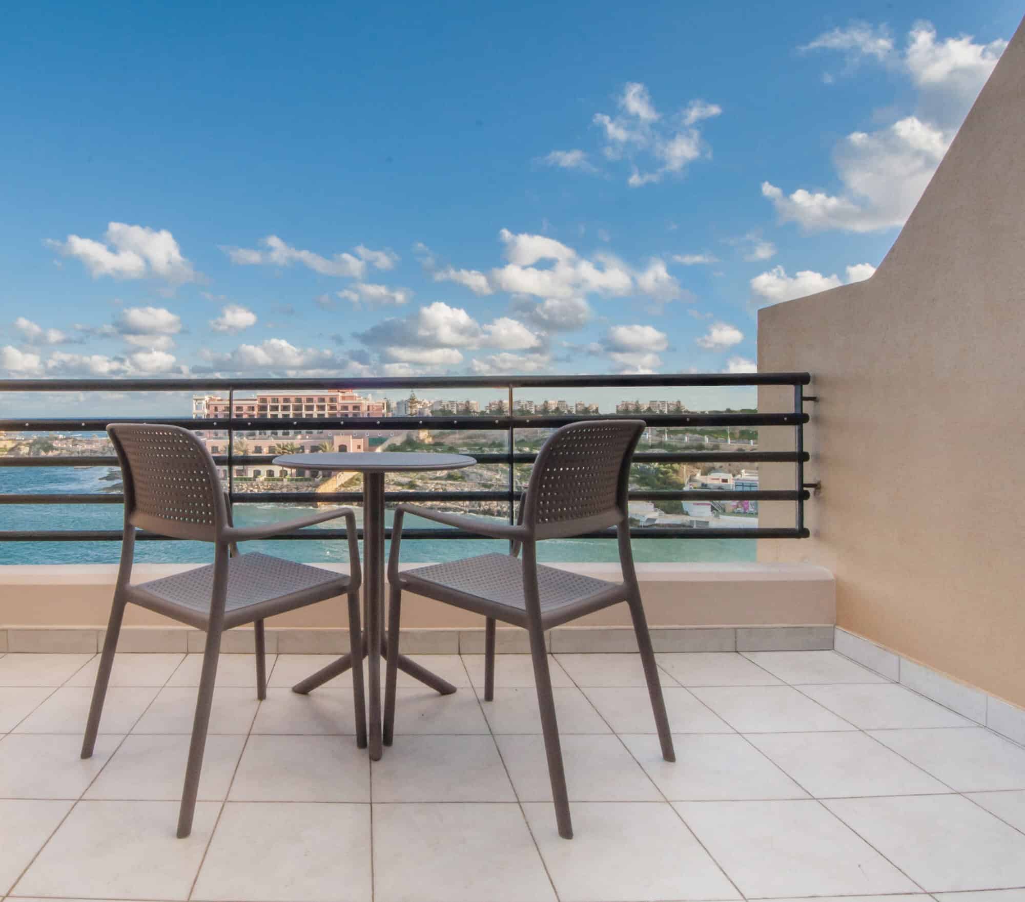 A small outdoor terrace with two chairs and a round table overlooking a body of water and a distant cityscape under a blue sky with scattered clouds.