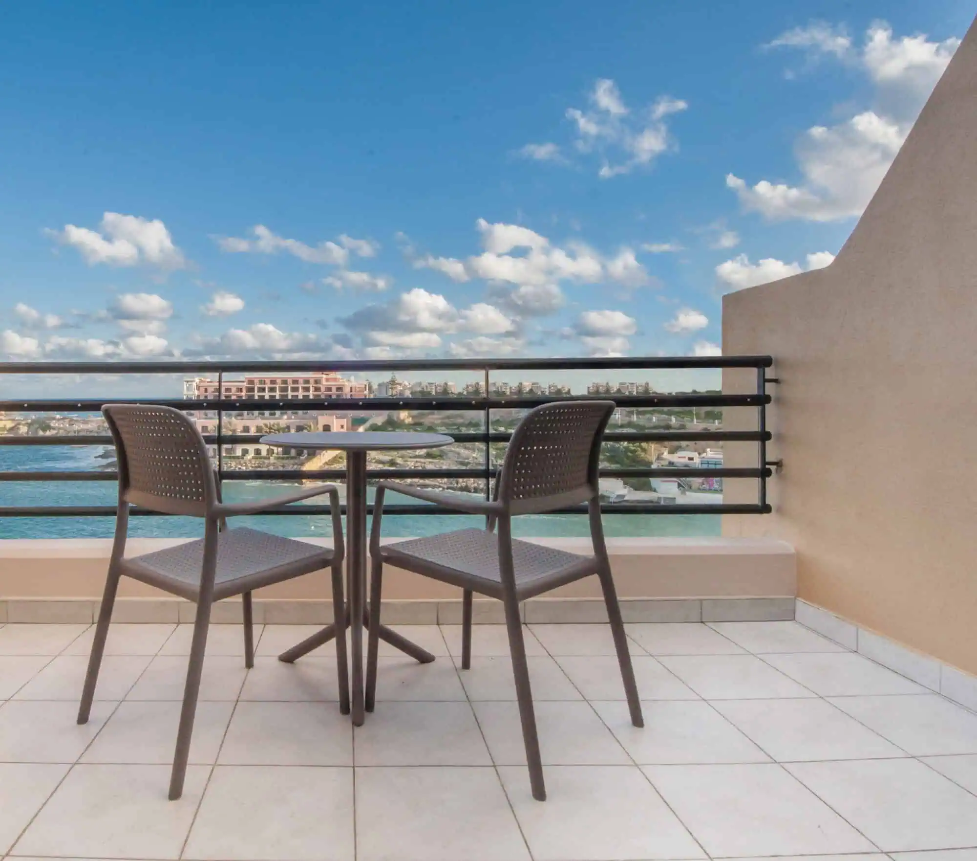 A small outdoor terrace with two chairs and a round table overlooking a body of water and a distant cityscape under a blue sky with scattered clouds.