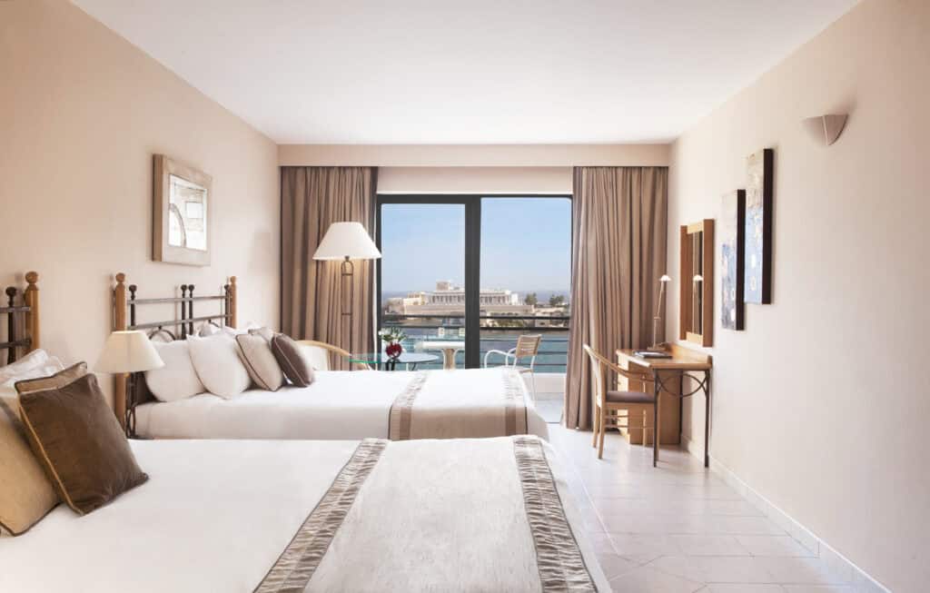 Hotel room featuring two double beds, a desk with a chair, and a large window offering a view of a waterfront and distant buildings. Neutral color scheme with beige and brown accents.