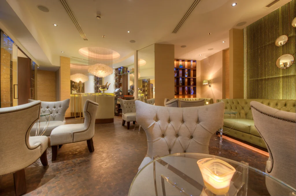 A modern, well-lit lounge with elegant tufted chairs, glass tables, a candle, and a bar area. The space features warm lighting, art on the walls, and a mix of contemporary decor elements.