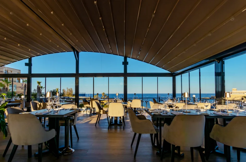 A modern restaurant interior with large windows overlooking the sea, set with white chairs and tables prepared for dining.