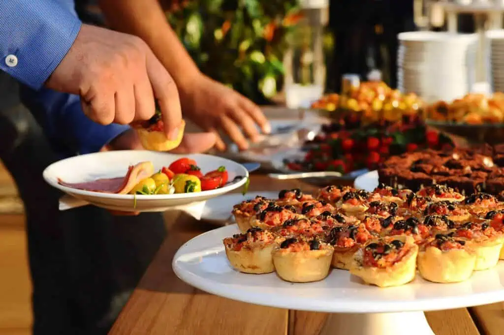 A person is serving themselves mini quiches from a platter at a buffet. Nearby are sliced meats, vegetables, and assorted desserts.