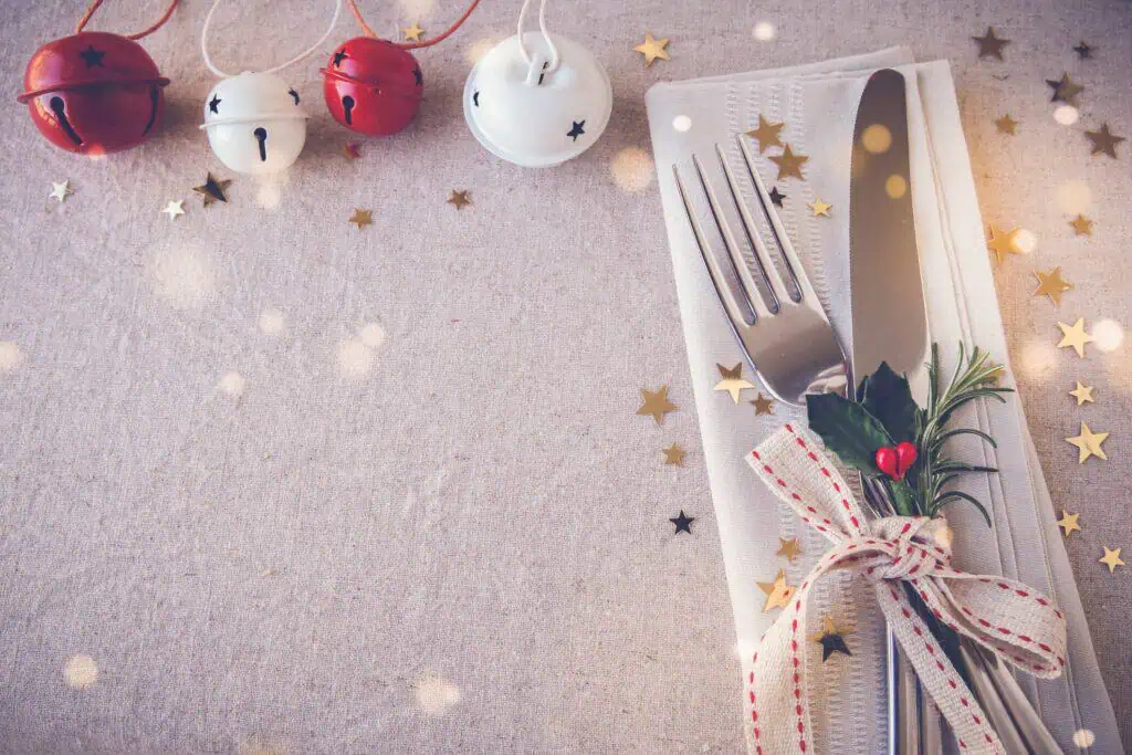 Festive table setting with a fork and knife on a napkin, decorated with a ribbon and greenery, next to scattered golden stars and red and white bells.