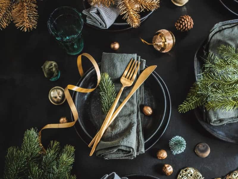 Festive table setting with a black plate, gray napkin, gold cutlery, and decorative pine branches on a black table. Gold ribbon accents and pinecones are scattered around.