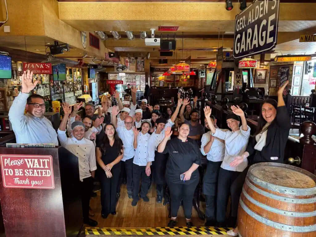 A group of restaurant staff, including chefs and servers, smiling and waving inside a rustic-themed restaurant with a "Please Wait to be Seated" sign and a "Geo. Alden's Garage" sign overhead.