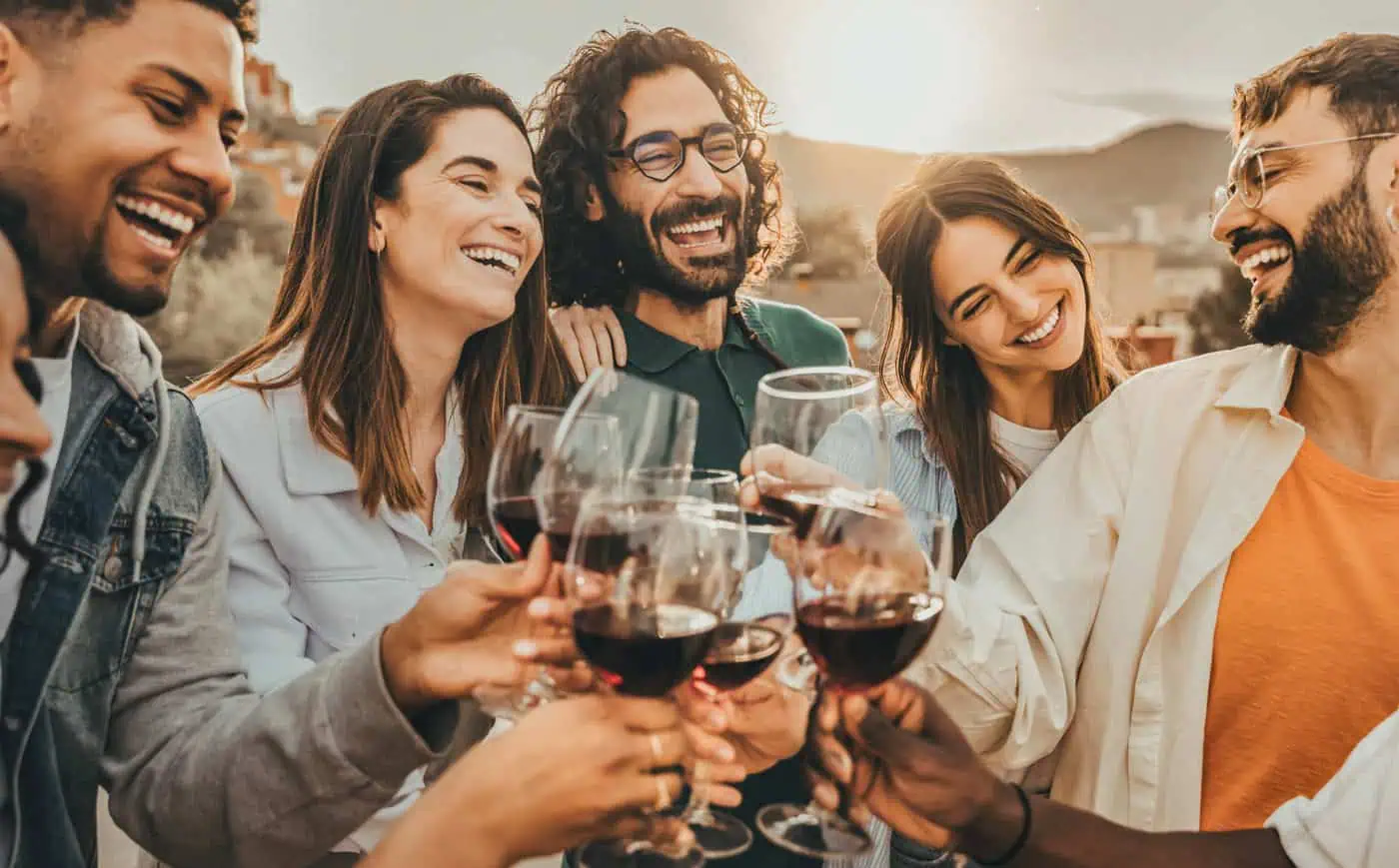 A group of six people smiling and clinking wine glasses outside during sunset.