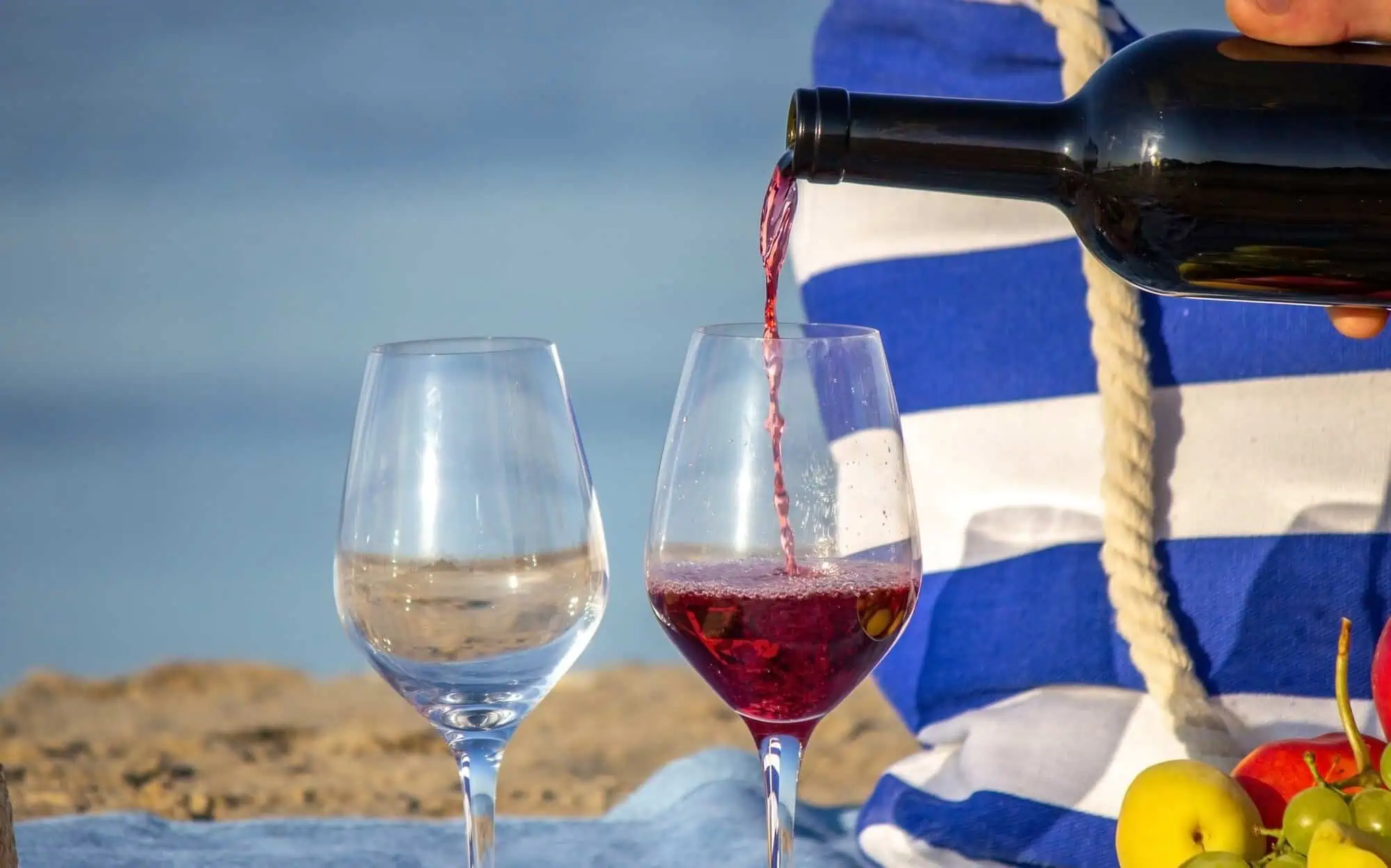 Red wine being poured into a glass next to another glass of clear liquid, with a blue and white striped bag and fruit on a beach background.