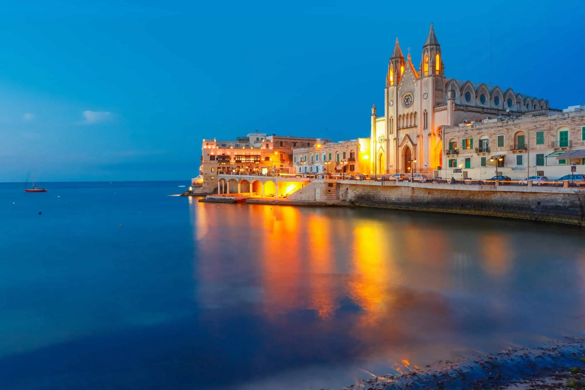 View of a lit-up church and buildings along a waterfront during twilight, with calm water reflecting the lights.