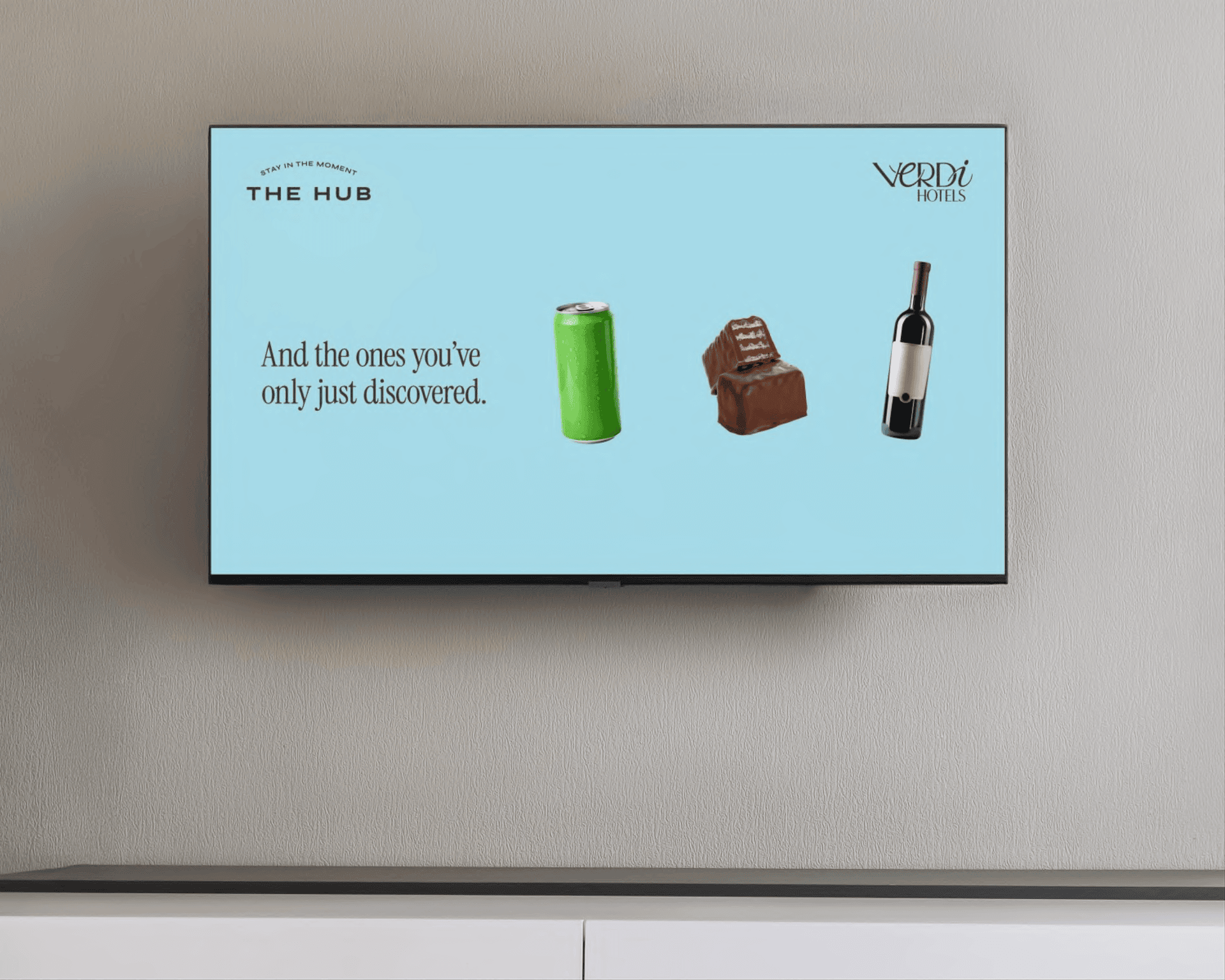 A wall-mounted TV screen displays an advertisement featuring a green beverage can, a chocolate bar, and a wine bottle against a light blue background with text promoting Verdi Hotels.