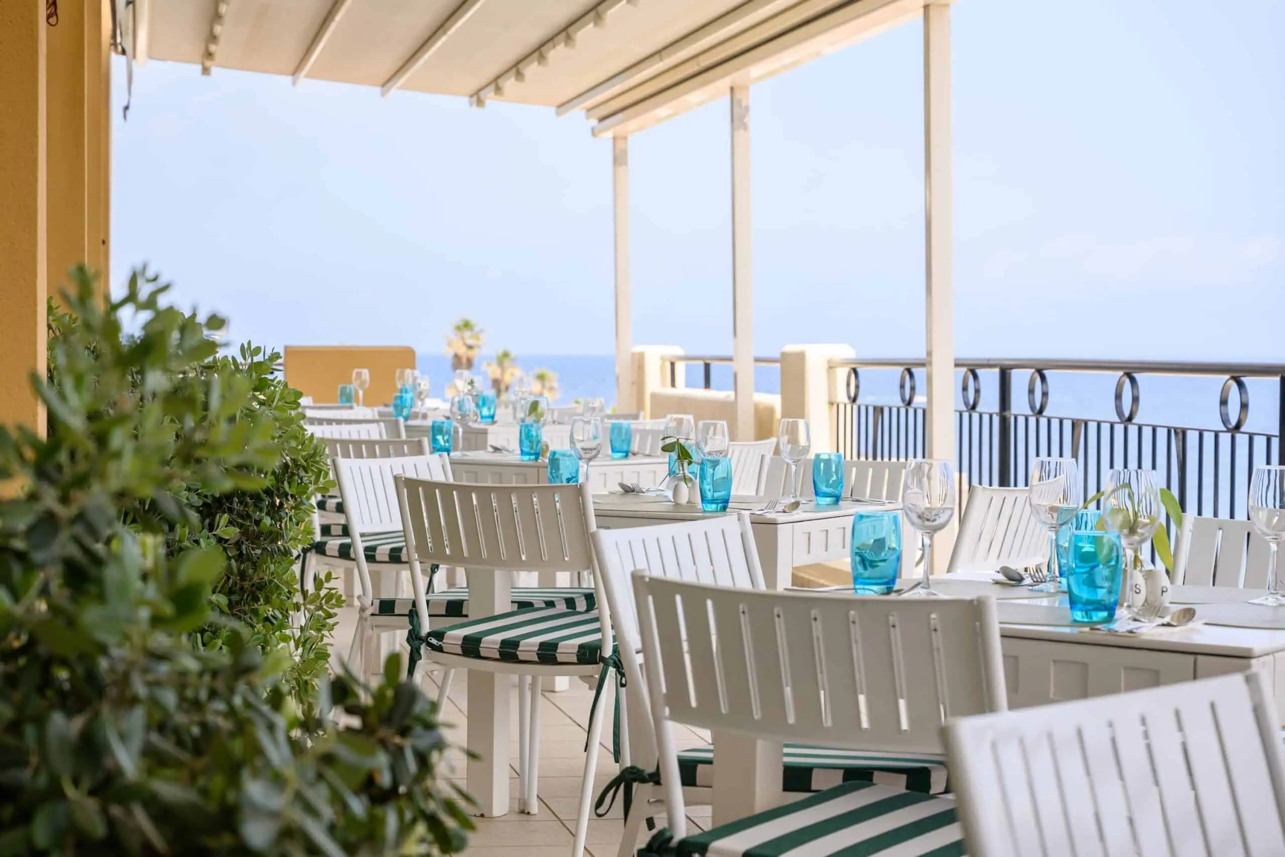 Outdoor restaurant terrace with white tables and chairs, striped seat cushions, blue glasses, and sea view in the background under a covered canopy.