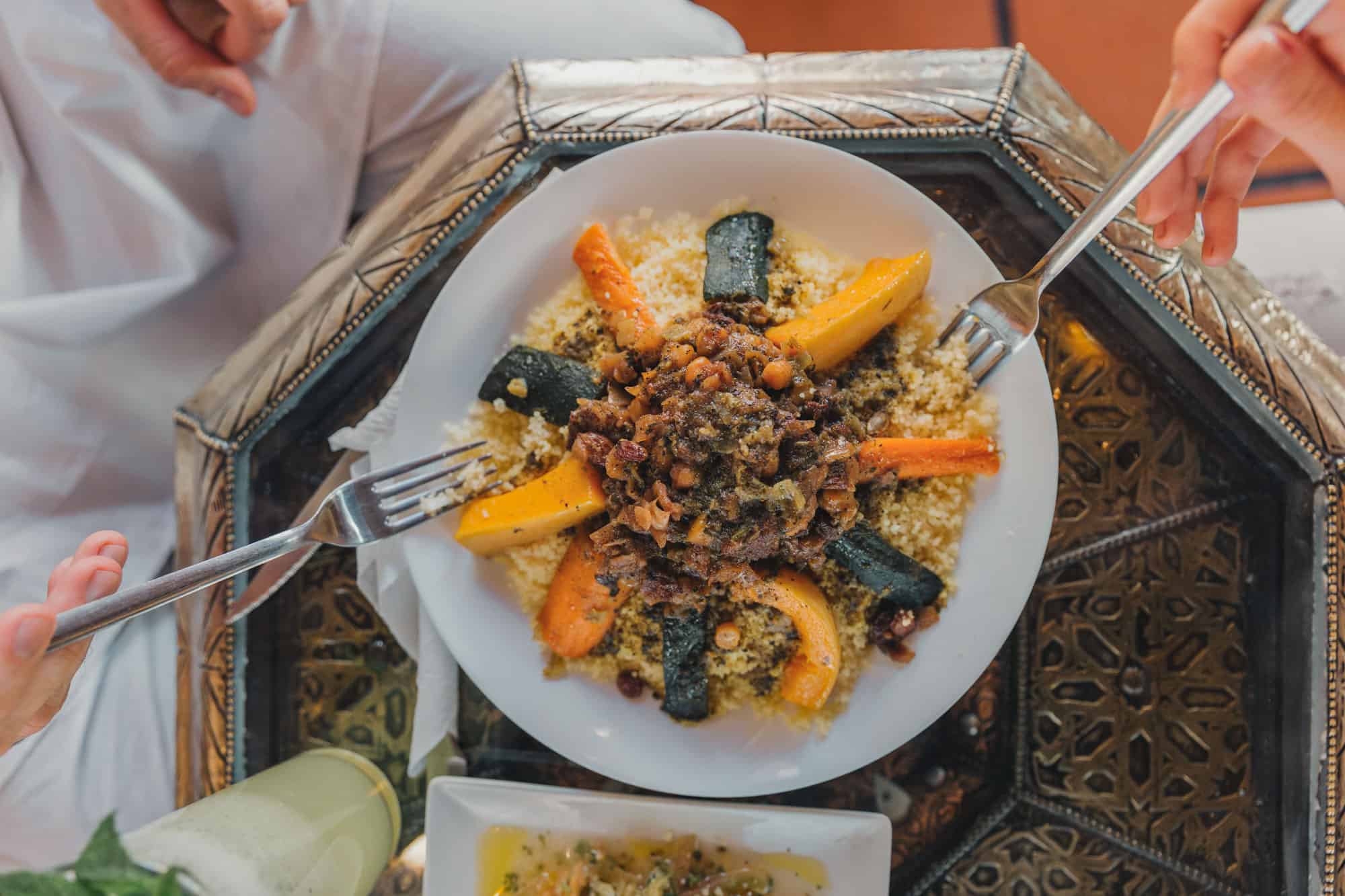 Plate of couscous topped with vegetables and meat, with two hands holding forks, on a decorative table.