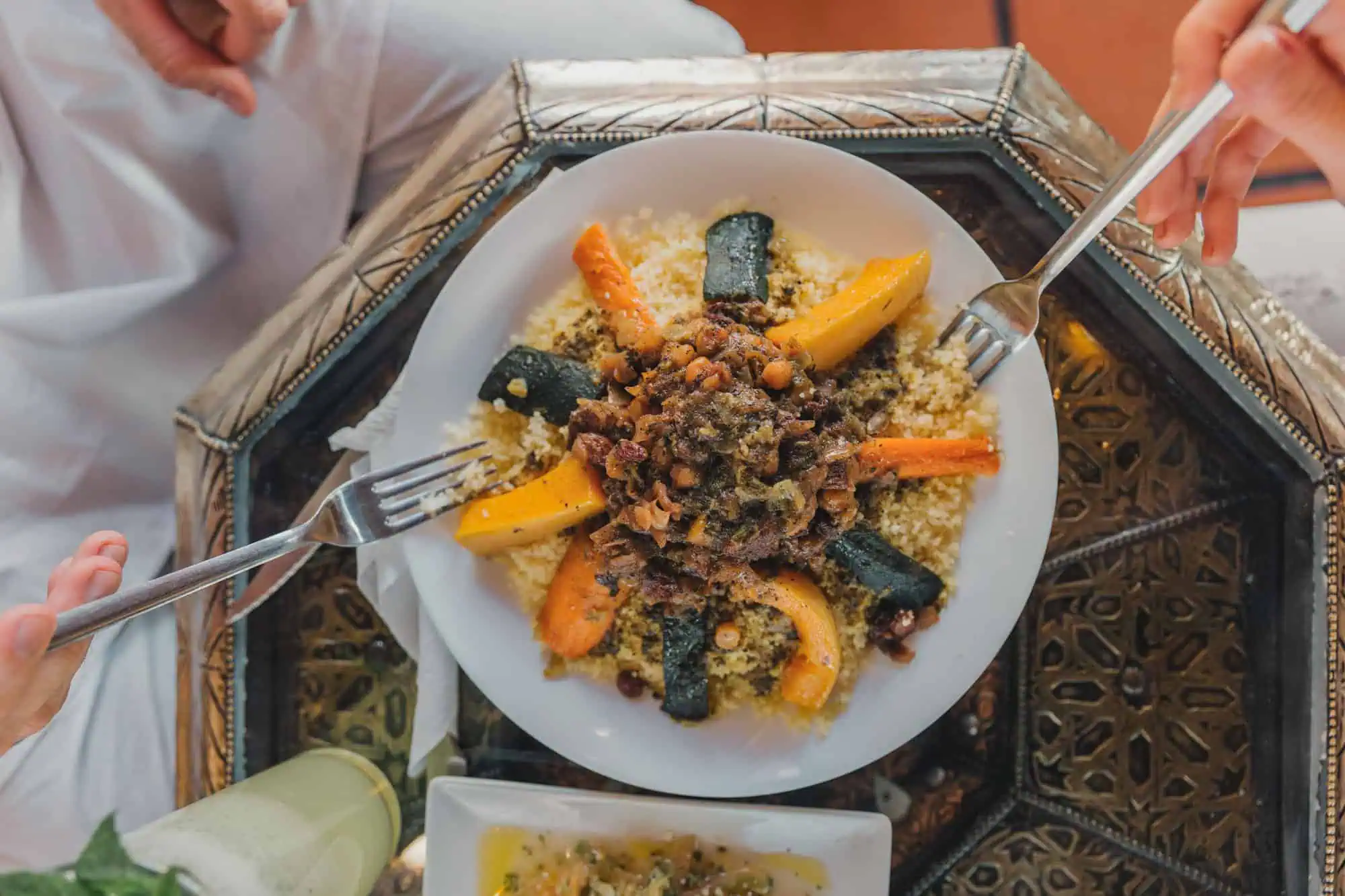 Plate of couscous topped with vegetables and meat, with two hands holding forks, on a decorative table.