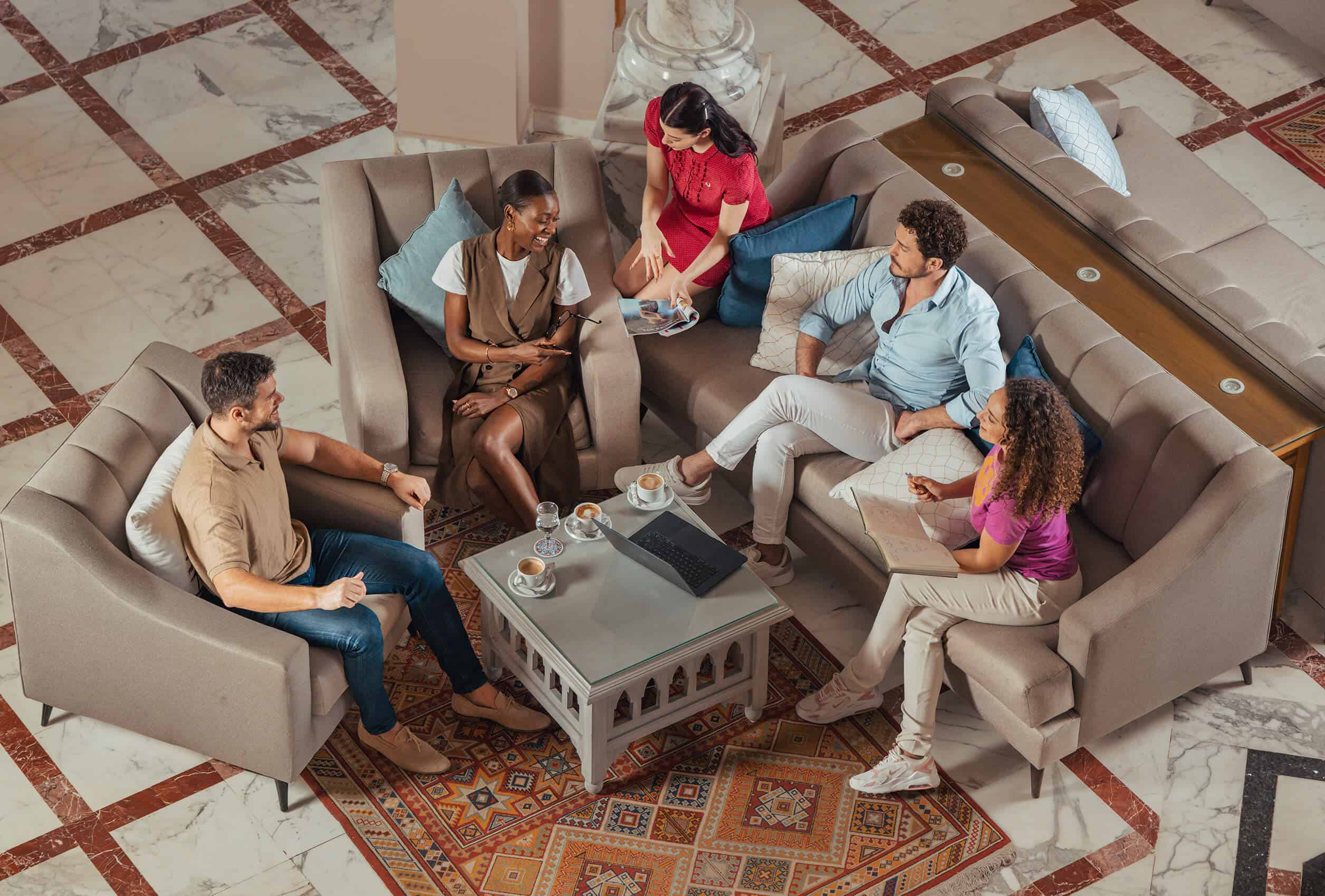 Five people in a meeting sit on sofas around a coffee table with laptops and cups, in a room with ornate rugs and tiled flooring.