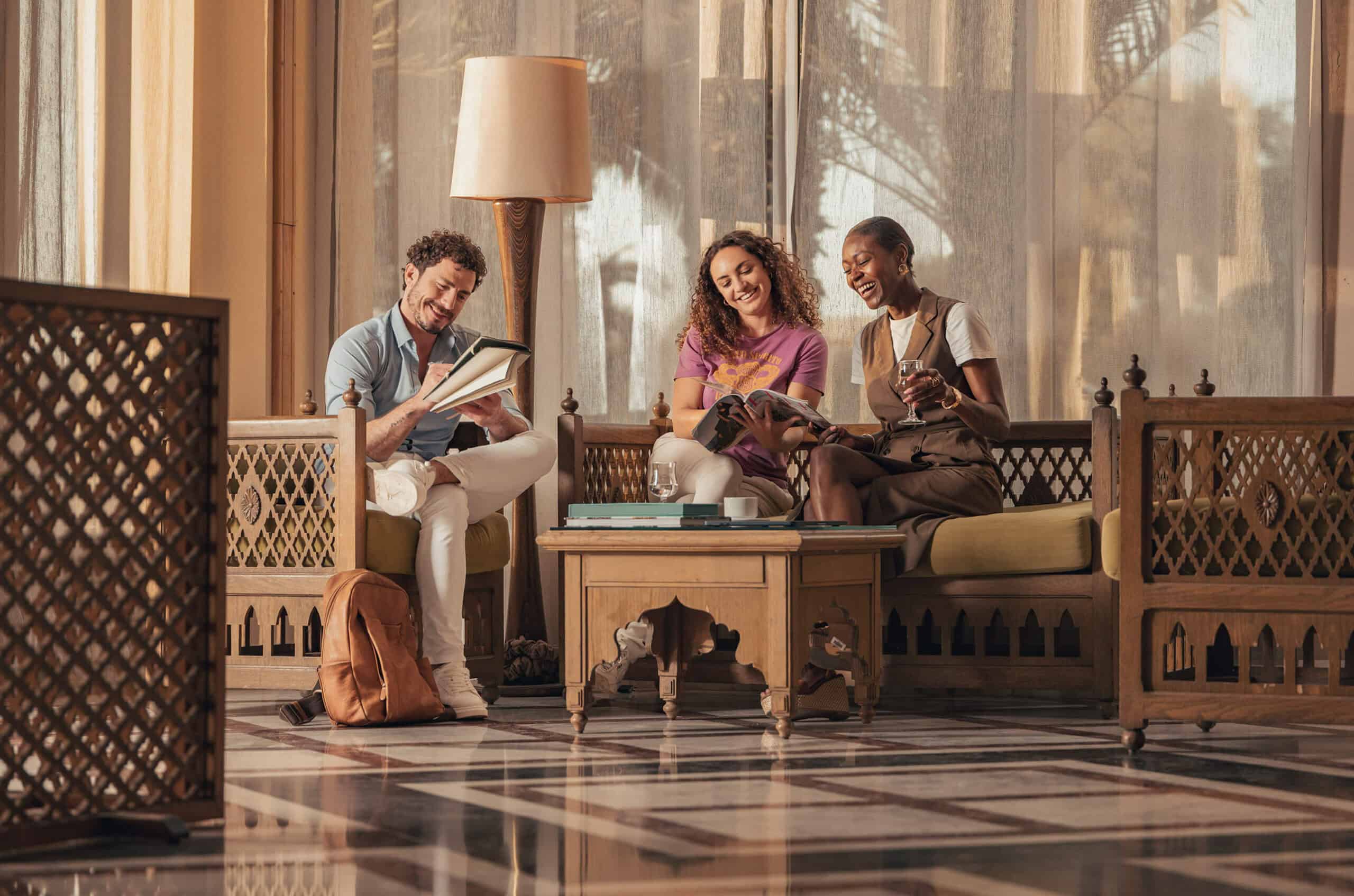 Three people sit on a wooden bench in a sunlit room, engaging in reading and conversation. A backpack is on the floor beside them.