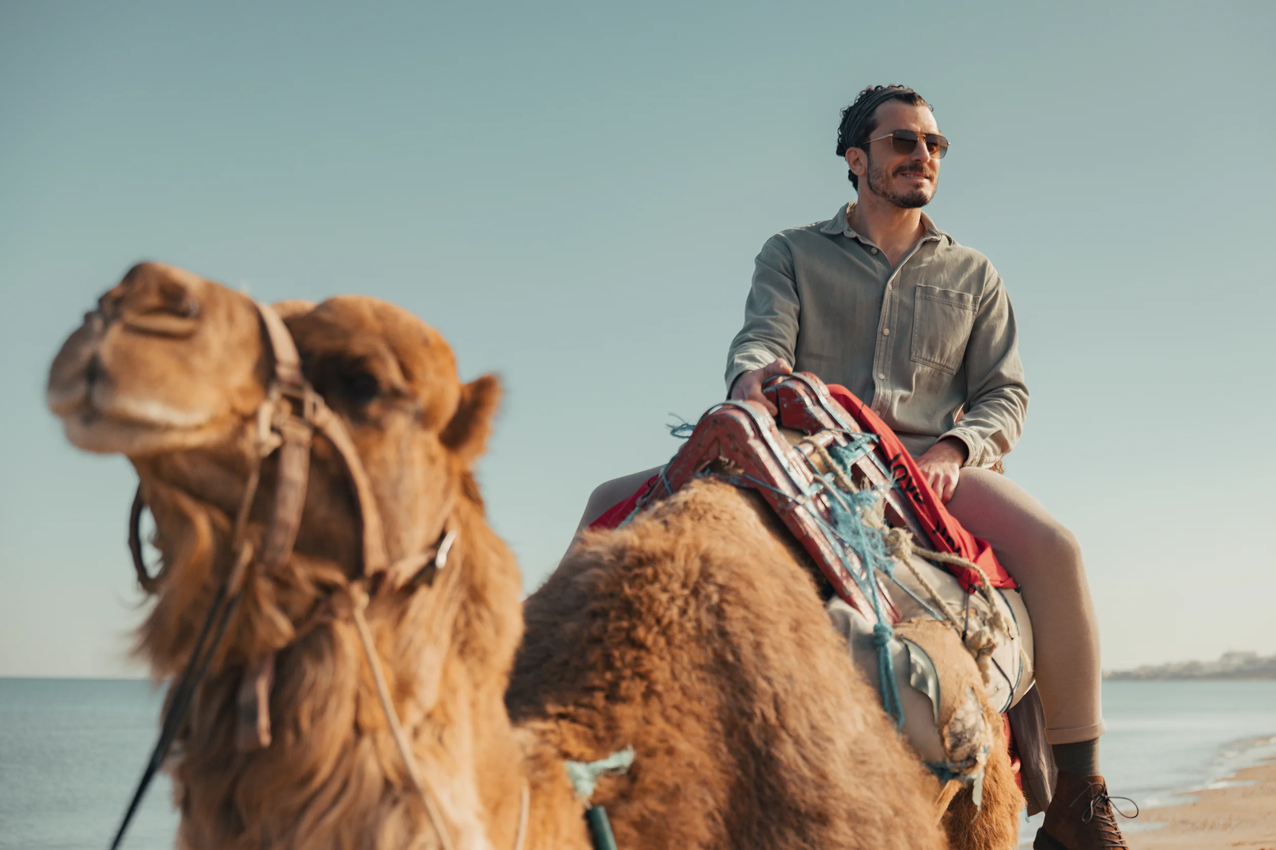 Man wearing sunglasses riding a camel on a beach with a clear sky.
