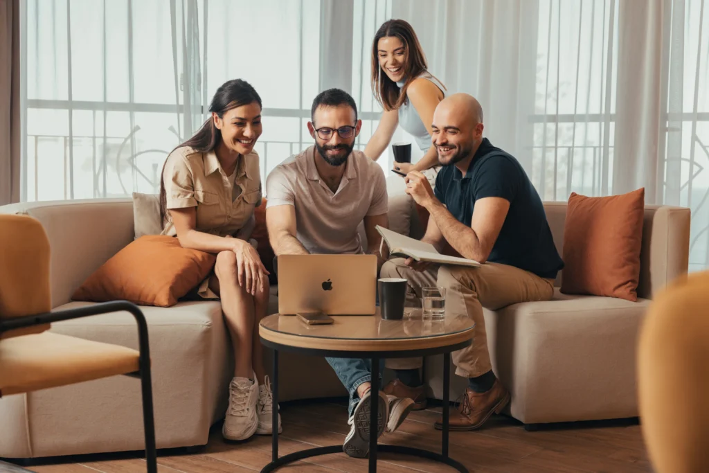 Four people sitting on couches, collaborating over a laptop with documents and beverages on a round table.