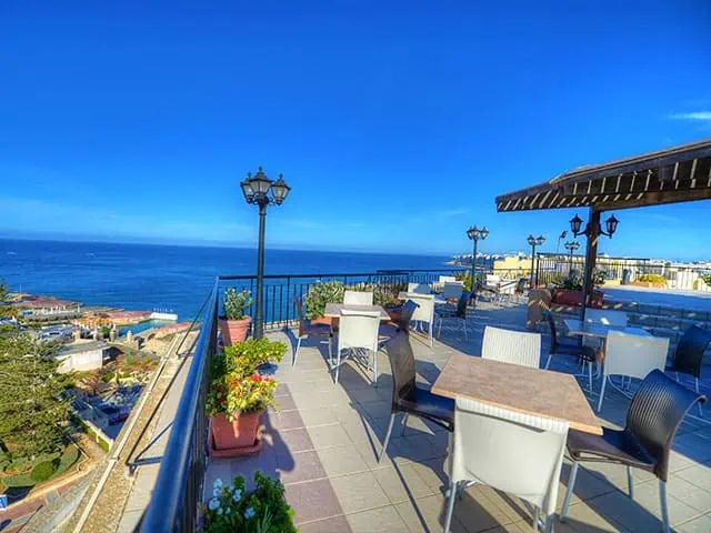 Rooftop terrace with tables and chairs overlooking a coastal view. Clear blue sky and ocean are visible in the background.
