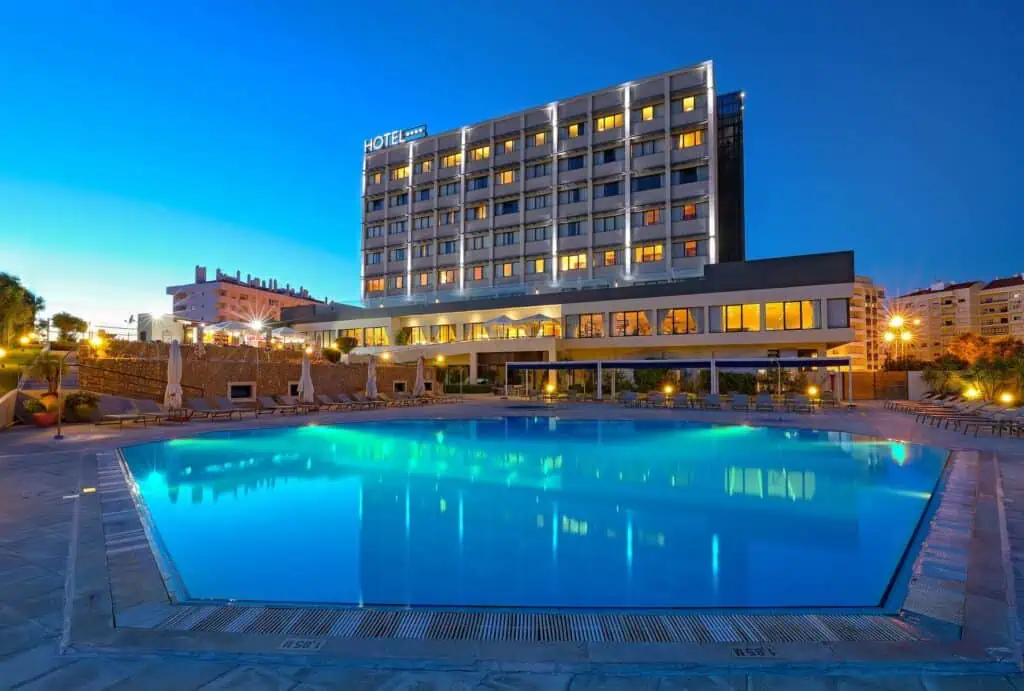 Modern multi-story hotel building at dusk with illuminated windows, situated behind a large, clear outdoor swimming pool surrounded by loungers and umbrellas.
