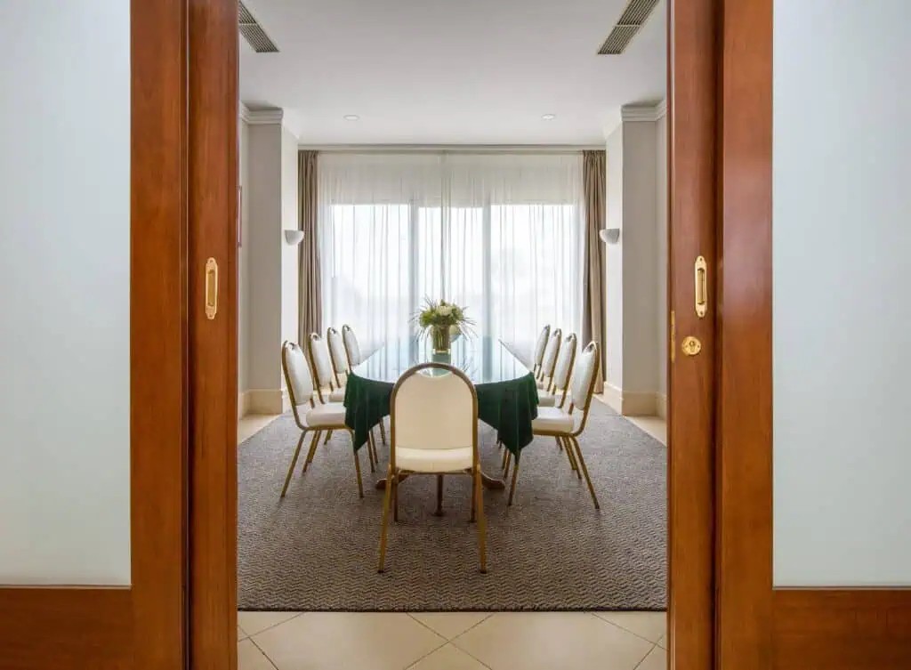 A dining room with a glass table, ten white chairs, a green tablecloth, and a flower centerpiece, viewed through open wooden doors.