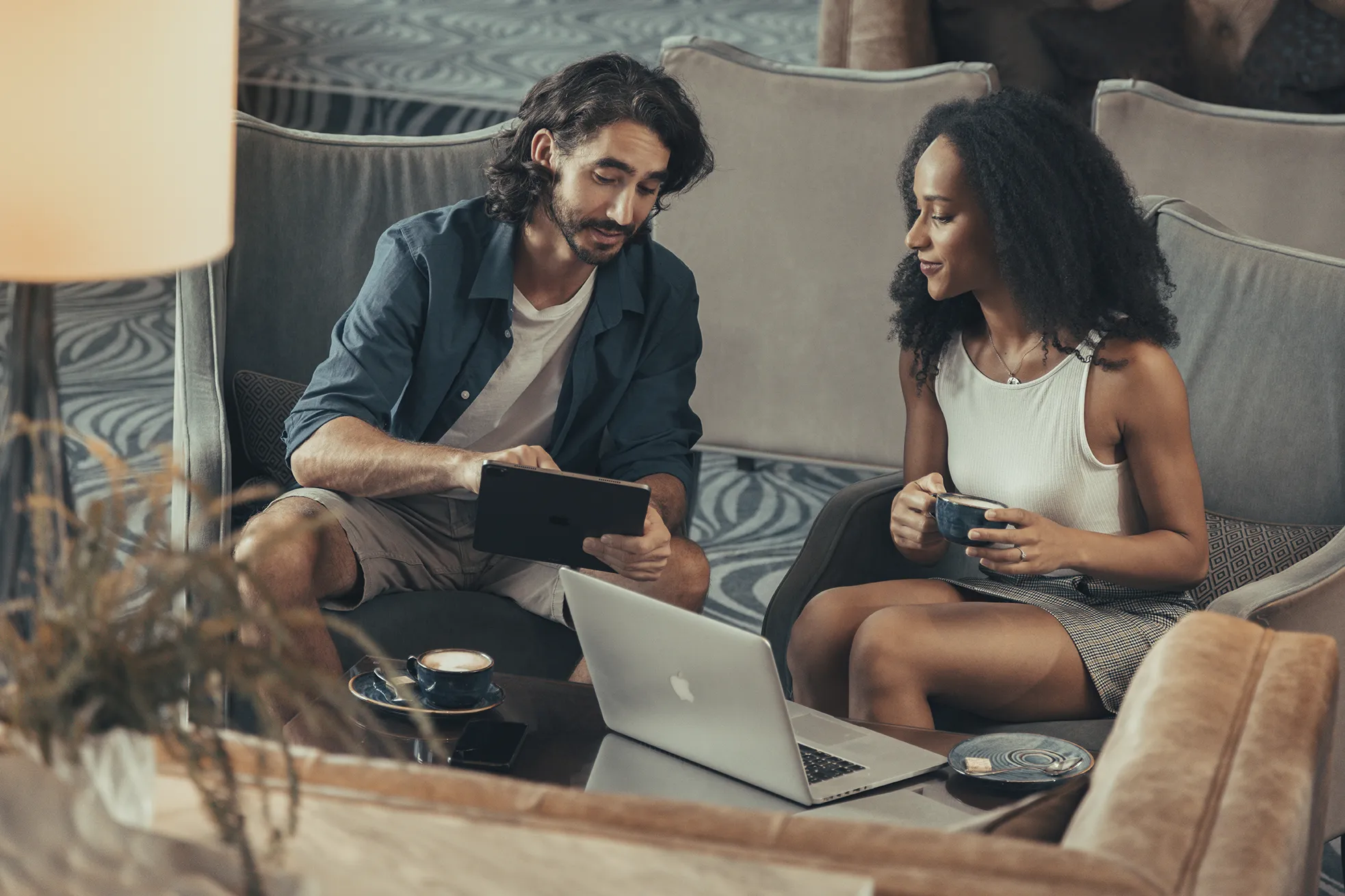 Two people sitting on couches, one holding a tablet and the other holding a cup, having a discussion, with a laptop and coffee cups on the table in front of them.