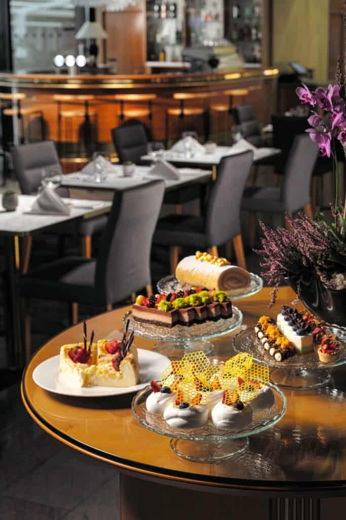 A variety of colorful desserts are displayed on glass plates atop a round wooden table in a restaurant with neatly set tables and modern chairs in the background.