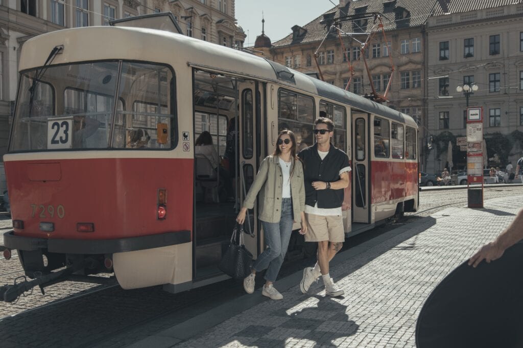 A young couple steps off a vintage red and cream tram at a city square on a sunny day, surrounded by historic buildings.