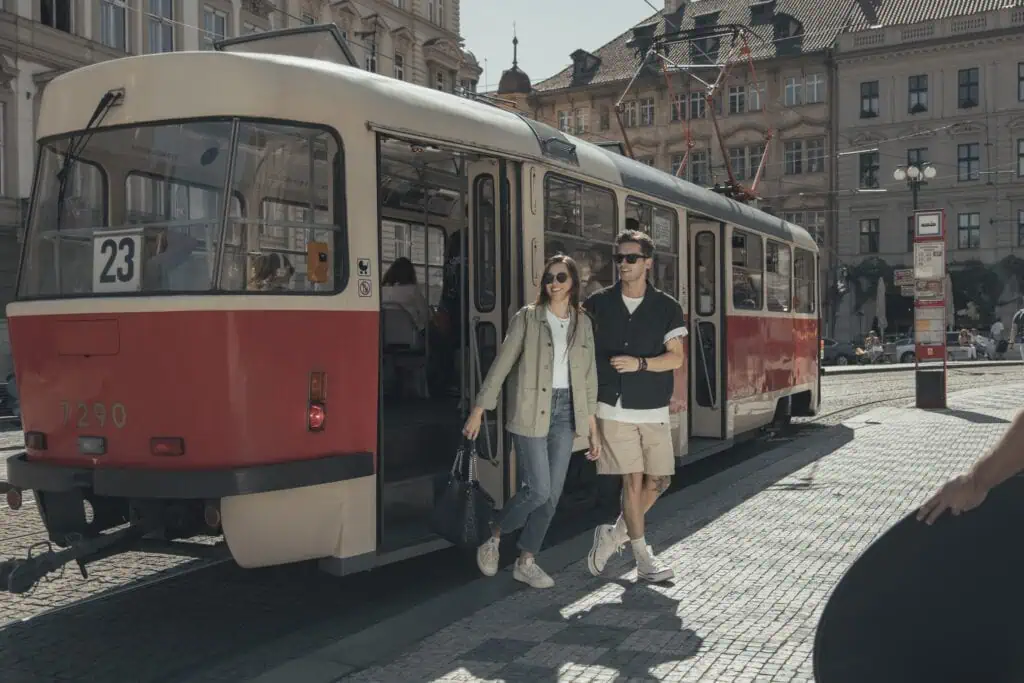 A young couple steps off a vintage red and cream tram at a city square on a sunny day, surrounded by historic buildings.