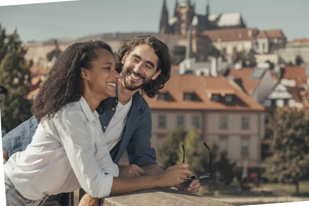 A smiling couple stands outdoors on a sunny day, leaning on a stone railing, with a historic cityscape featuring red rooftops and towers in the background.